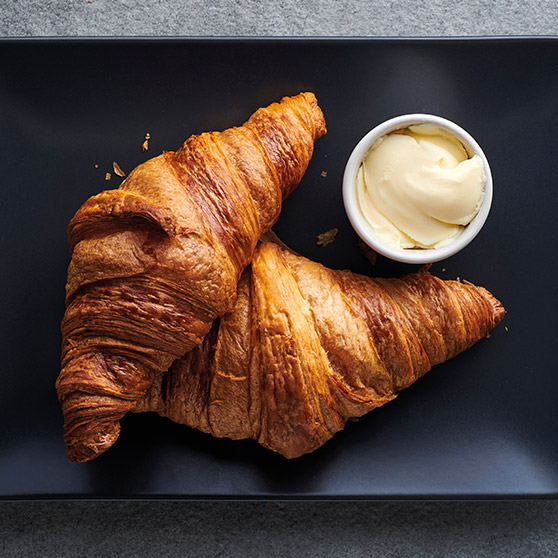 Due croissant dorati appena sfornati su un piatto nero con una ciotola di burro – preparati con il CHEF'S COMBI di HOBART per una perfetta doratura e consistenza.