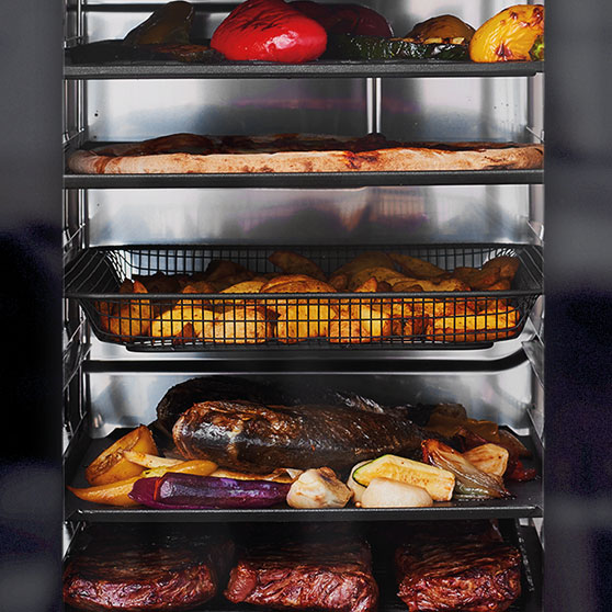 Vista interna di un forno CHEF'S COMBI di HOBART con più ripiani – con verdure grigliate, pizza, patate al forno, arrosto con verdure e bistecche, tutto cotto in modo uniforme e simultaneo.