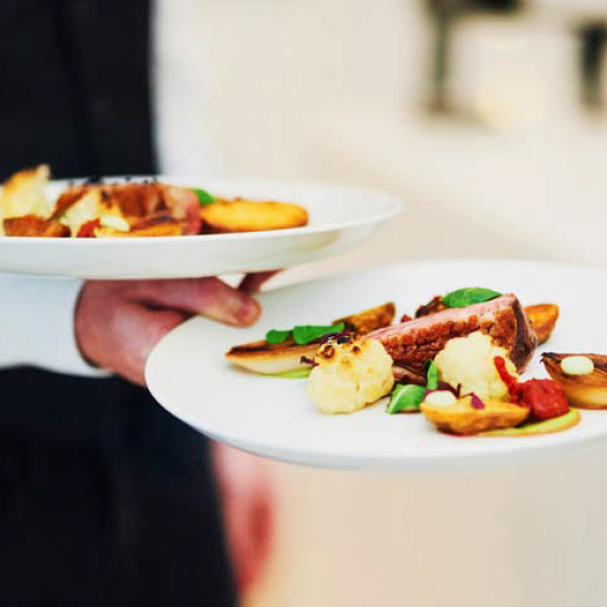 Una persona tiene due piatti elegantemente impiattati con carne, verdure arrosto e guarnizioni raffinate – preparati con il CHEF'S COMBI di HOBART per soddisfare gli standard più elevati della cucina professionale.