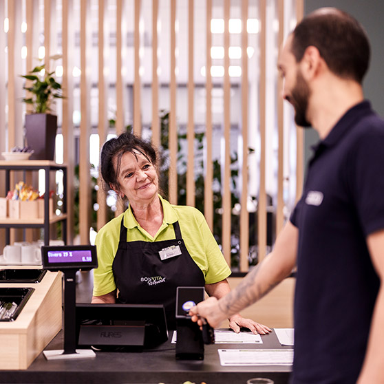 Pagamento in una mensa aziendale moderna: Un cliente effettua un pagamento contactless con carta su un terminale elettronico. L’importo è visibile su display digitale. Sullo sfondo listelli in legno e piante creano un’atmosfera accogliente.