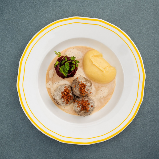 Piatto bianco con bordo giallo su tavolo grigio, con polpette Königsberger in salsa cremosa chiara e contorno, servite in modo tradizionale.
