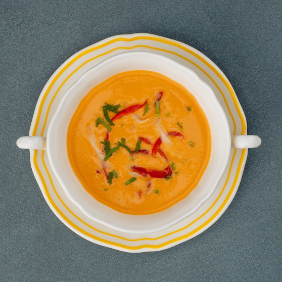 Tazza da zuppa bianca con bordo giallo su piatto, riempita con zuppa cremosa arancione. La tazza ha due manici ed è posizionata su un tavolo grigio.