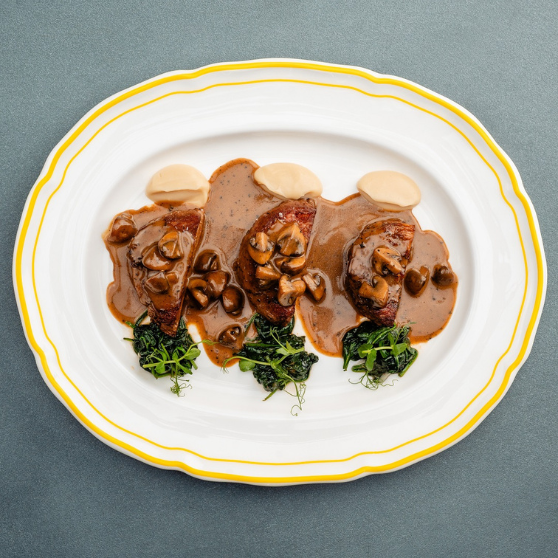 Piatto bianco con bordo giallo con tre pezzi di carne in salsa cremosa ai funghi, guarniti con funghi, purè chiaro e erbe verdi, servito in modo elegante su tavolo grigio.
