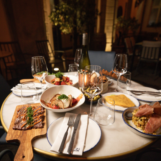Tavolo da ristorante all’aperto elegantemente apparecchiato con vari piatti, antipasti, pane, una bottiglia di vino e diversi bicchieri.