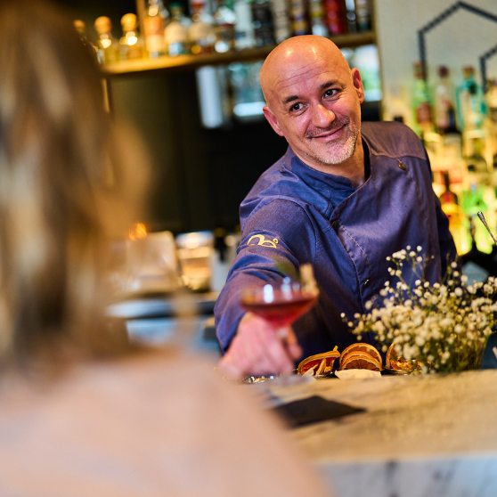 Barista che serve una bevanda a un ospite oltre il bancone, con bottiglie e scaffali del bar sullo sfondo.