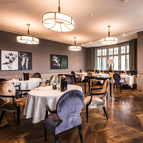 Interno elegante di un ristorante fine‑dining con tavoli rotondi apparecchiati in bianco, sedie imbottite, decorazioni artistiche e illuminazione calda.