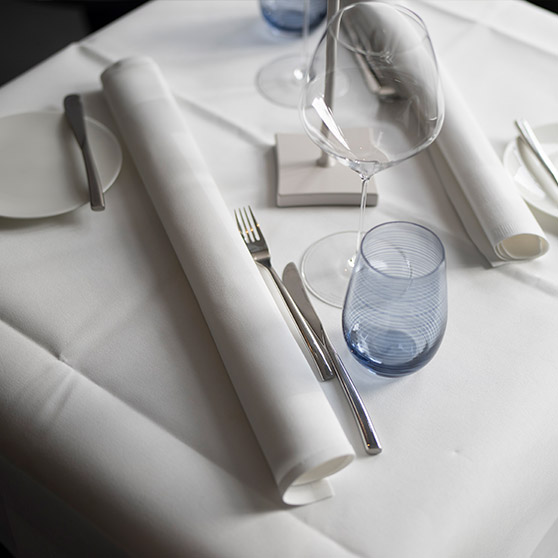 Tavolo da ristorante elegantemente apparecchiato con posate disposte con cura, calici da vino, tovaglioli arrotolati e bicchiere blu, creando un’atmosfera raffinata.